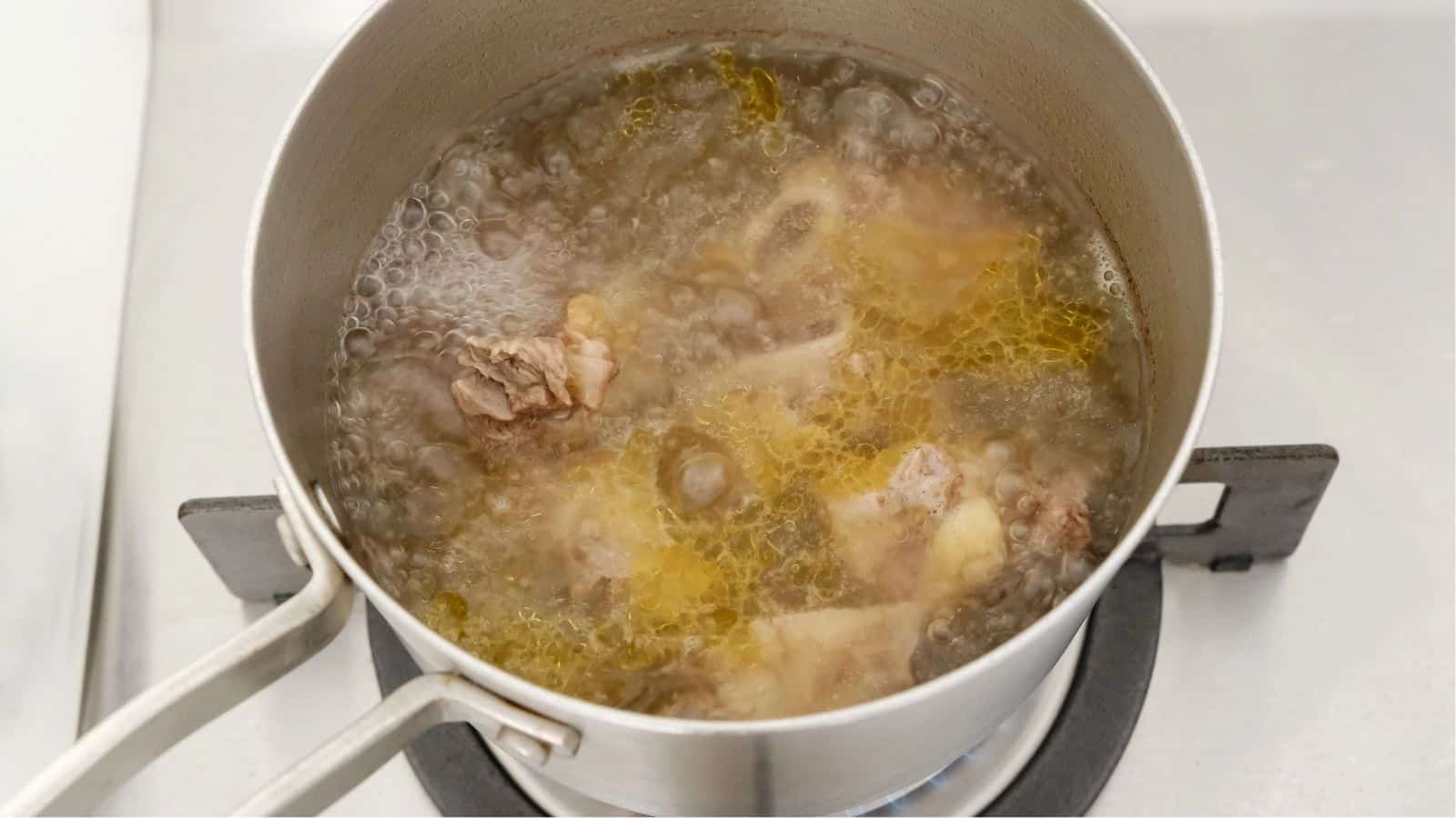 A metal pot filled with bones and meat is boiling on a gas stove, producing broth with bubbles and a layer of oil on the surface.