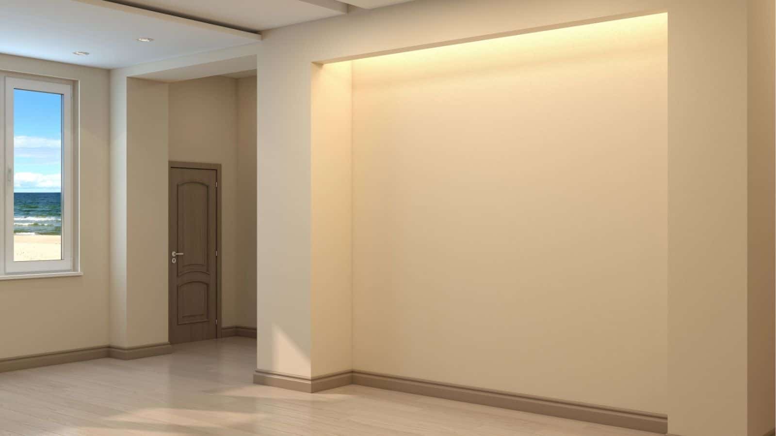 An empty beige room with light wood flooring, a window showing a beach view, a closed door, and a recessed alcove with overhead lighting.