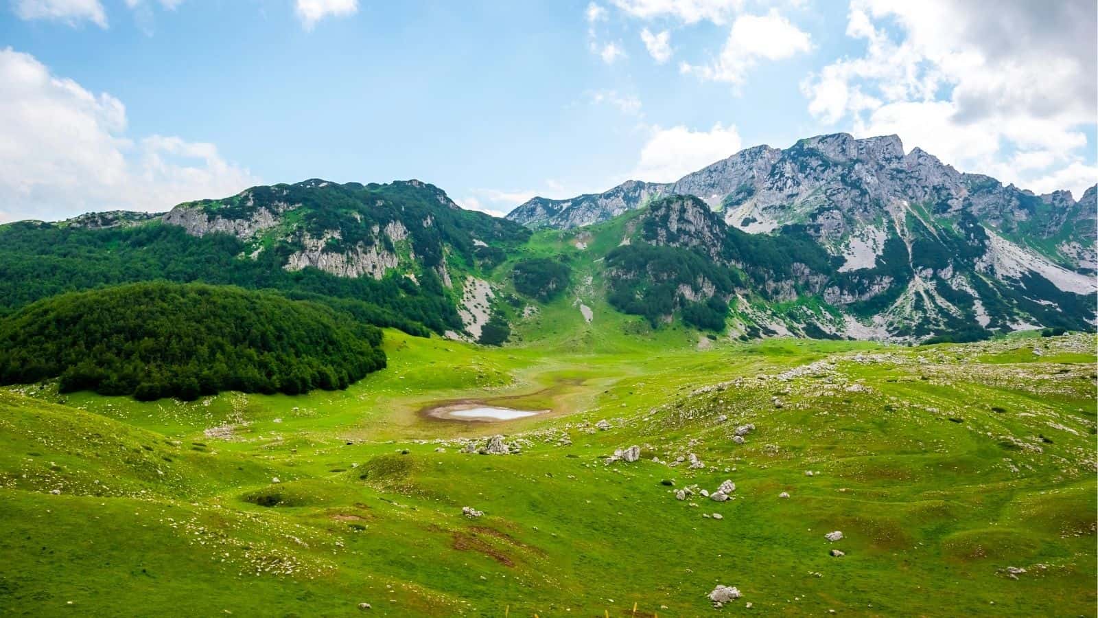 Green mountains and hills with rocky patches and a small pond in a valley under a partly cloudy sky.