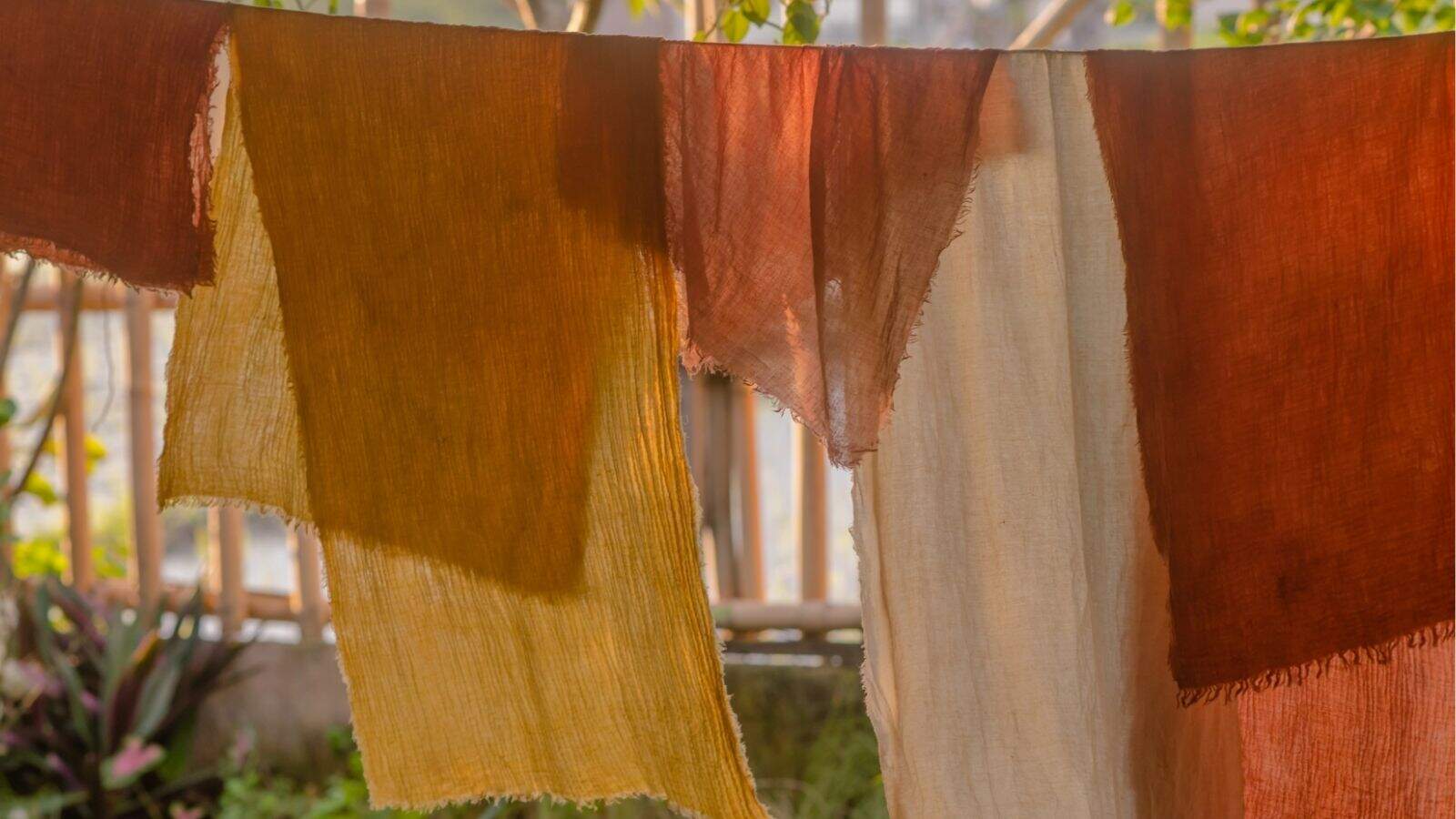 Several pieces of fabric in earthy tones, including brown, yellow, and cream, hang on a clothesline outdoors with plants visible in the background.