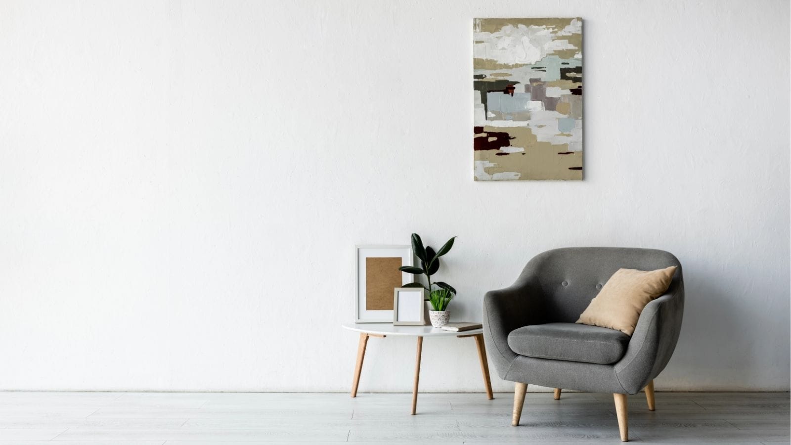 A modern living room with a gray armchair, beige pillow, small side table with framed art and a plant, and an abstract painting on a white wall.