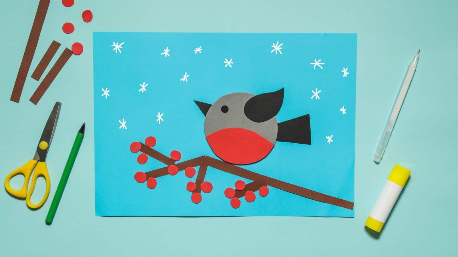 A paper craft of a bird on a branch with red berries and white snowflakes, surrounded by scissors, pencil, glue stick, and paper strips on a blue background.