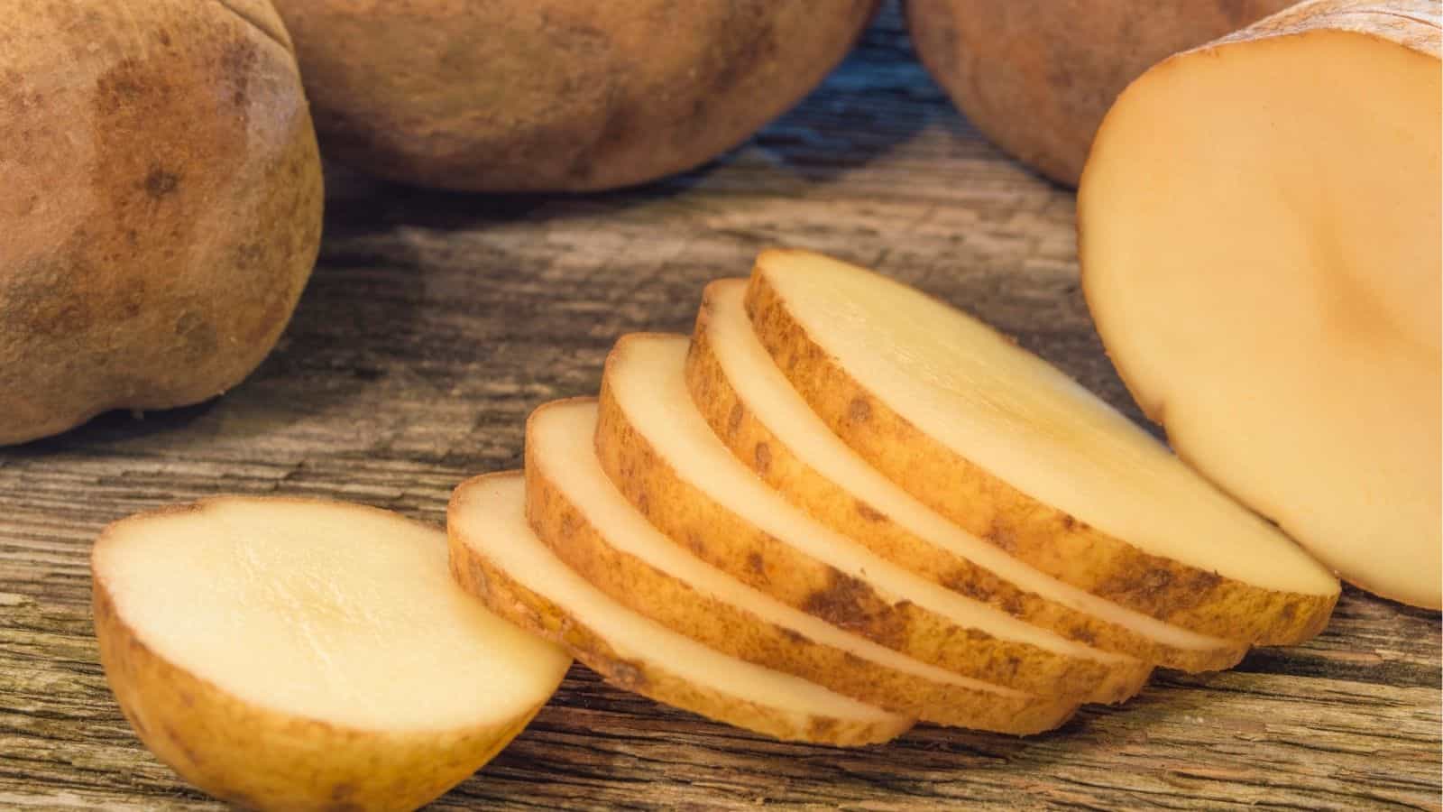 A close-up of a raw potato on a wooden surface, with one potato partially sliced into thin, even round pieces.