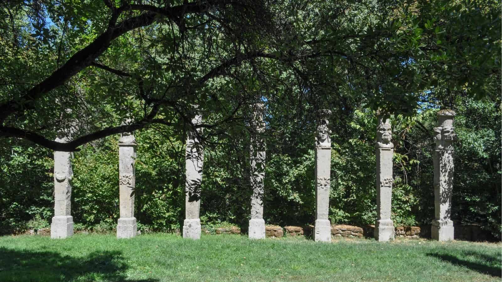 Six stone pillars with carved tops stand in a row on grass, surrounded by trees and greenery under a large overhanging tree branch.