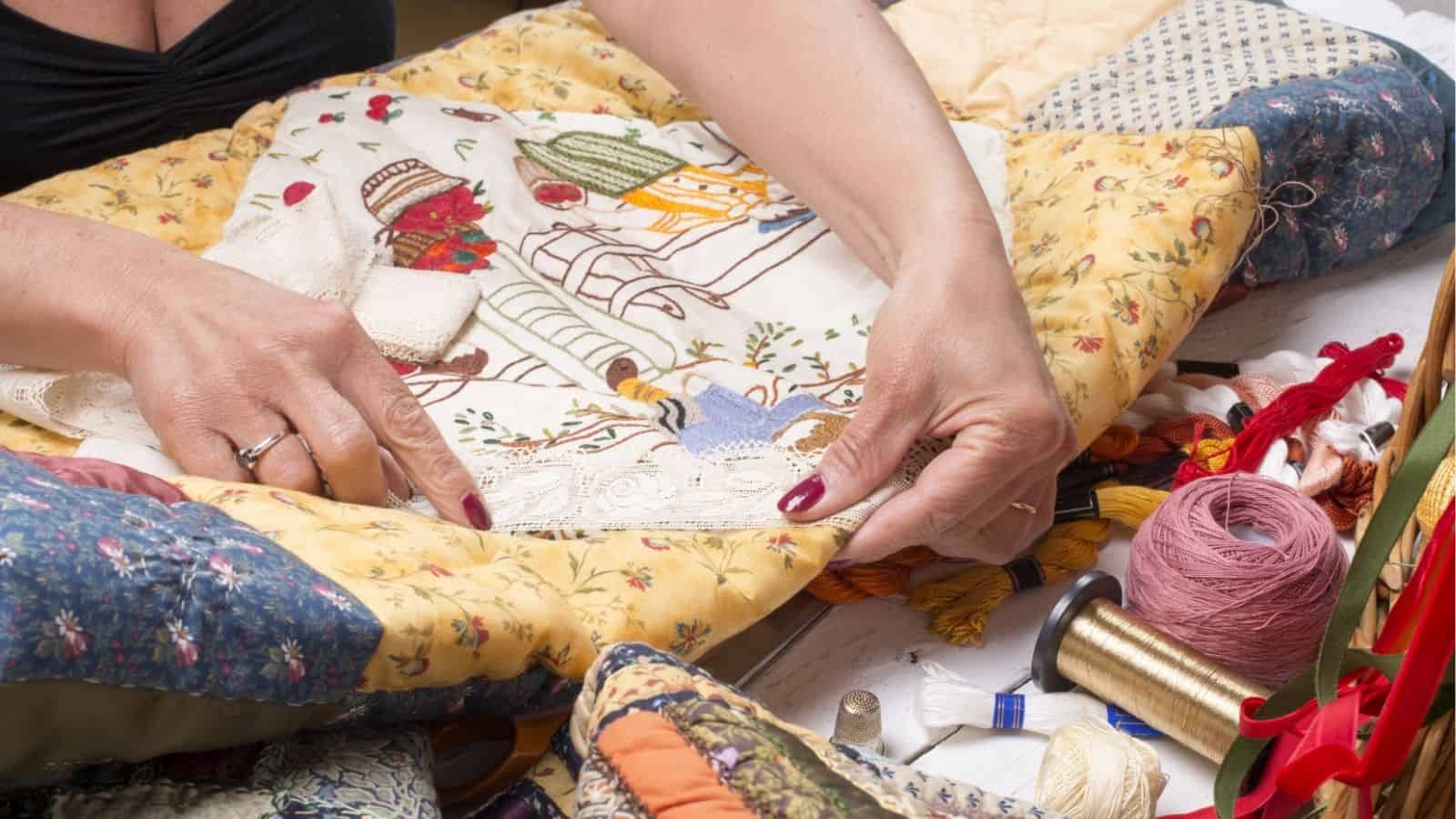 A person works on a colorful quilt using embroidery threads and sewing tools, with hands holding the fabric and various supplies scattered around.
