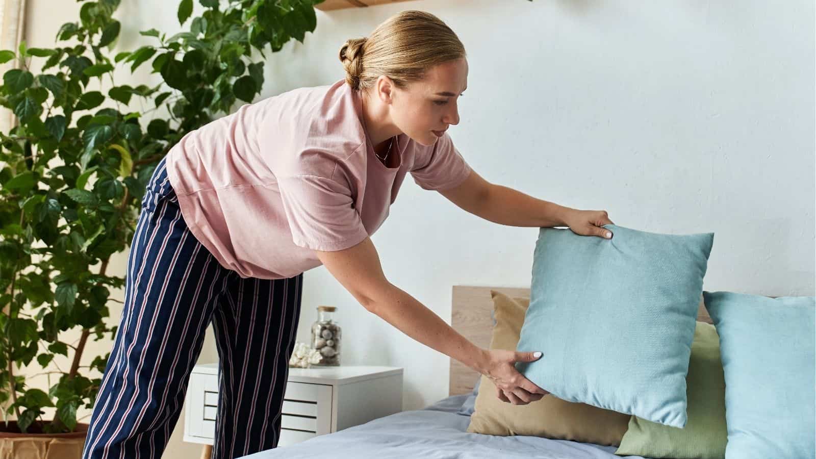 A woman in striped pajama pants arranges pillows on a neatly made bed in a bedroom with plants and a white nightstand.