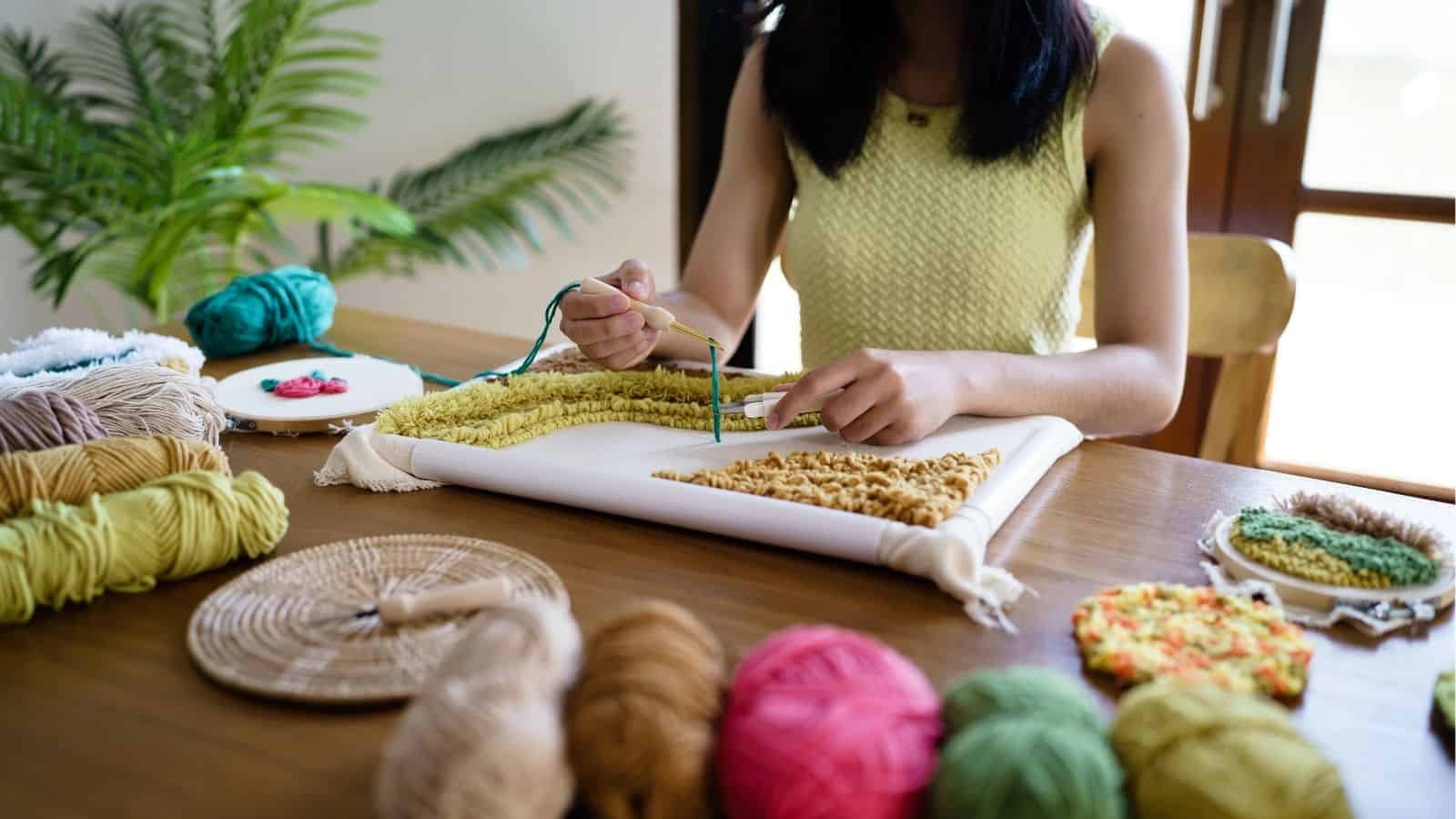 A person works on a yarn punch needle project at a wooden table, surrounded by colorful yarn, embroidery hoops, and craft materials.