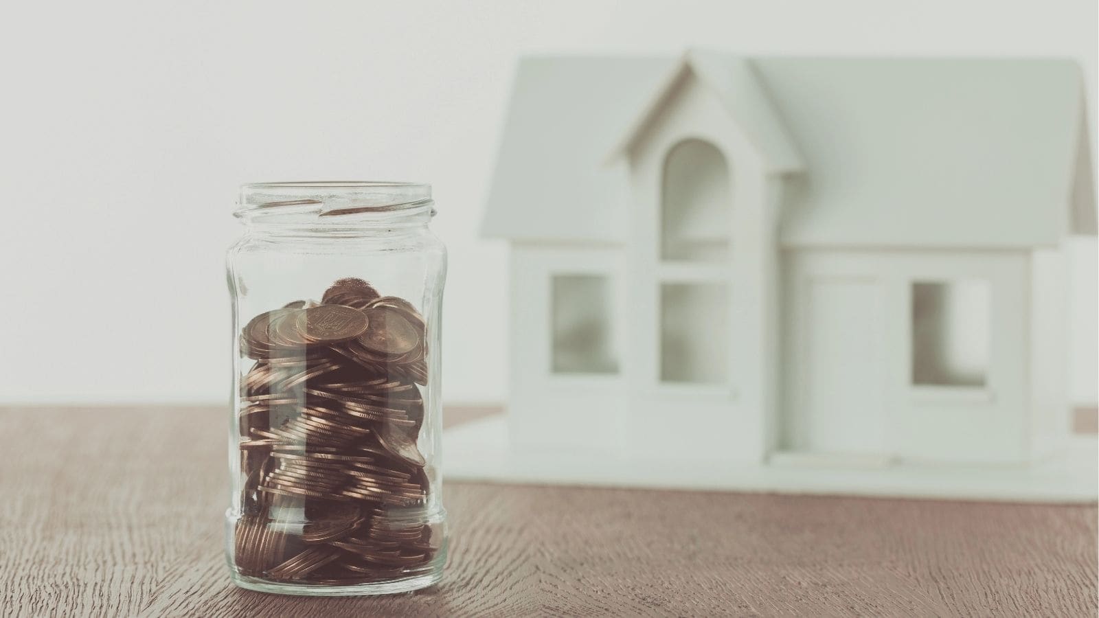 A glass jar filled with coins sits on a wooden surface in front of a blurred model of a white house.