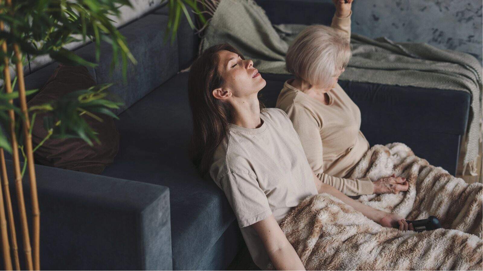 Two women sit on the floor leaning against a sofa, eyes closed, covered with a blanket, practicing meditation or relaxation.