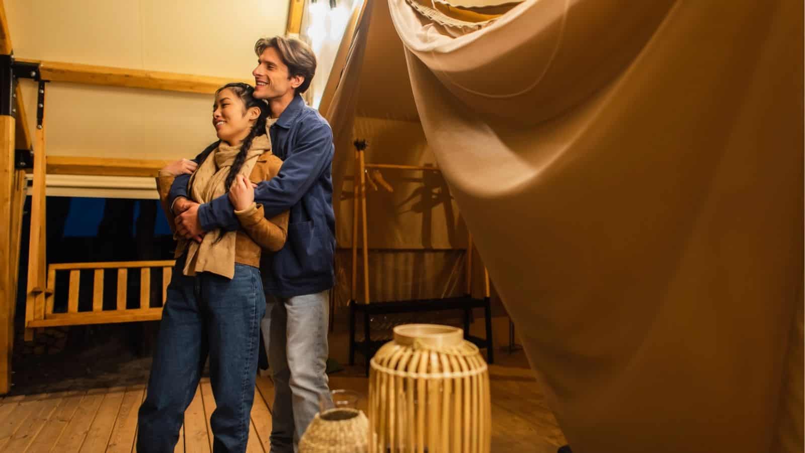 A couple stands inside a glamping tent, with the man hugging the woman from behind. Warm lighting and wooden decor create a cozy atmosphere.