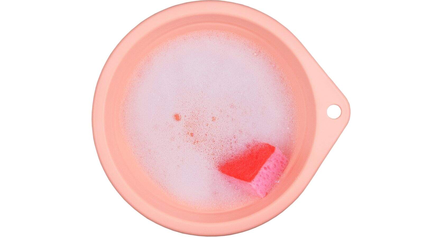 A pink sponge floats in a round, peach-colored bowl filled with soapy water and bubbles, viewed from above.