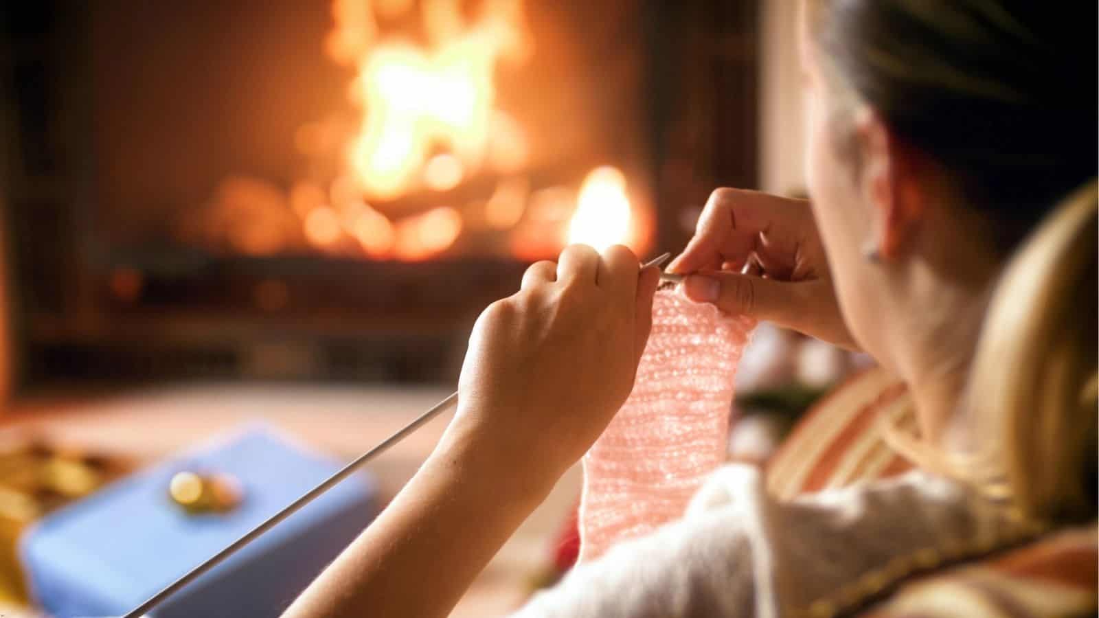 A person sits near a lit fireplace, knitting with pink yarn. A book and small object are visible on a table nearby.