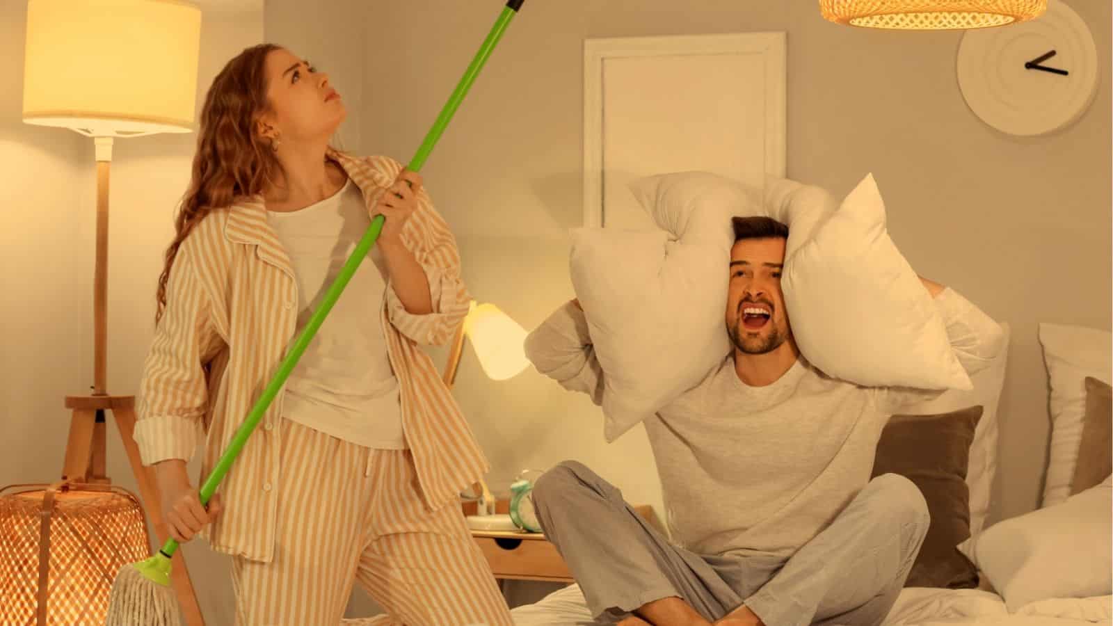 A woman in pajamas sings into a broom while a man sitting on a bed covers his ears with pillows, looking annoyed.