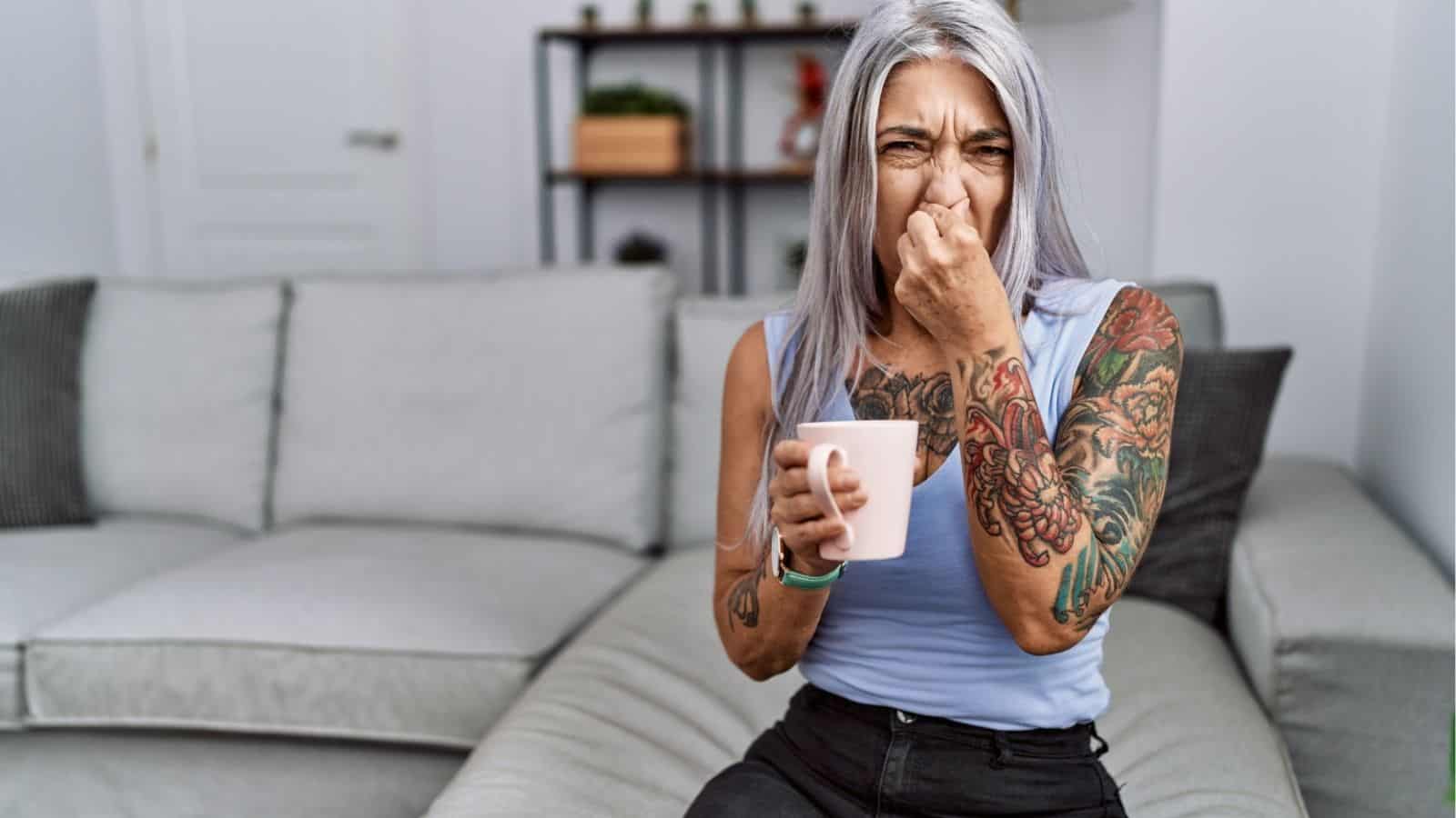 A woman with gray hair and tattoos sits on a couch, holding a mug and pinching her nose, appearing to react to an unpleasant smell.