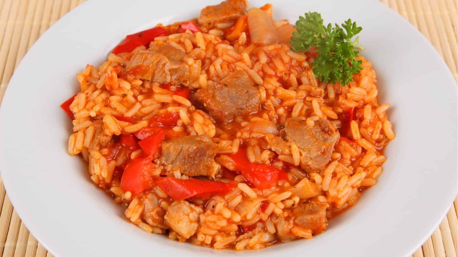 A white plate filled with rice, red bell peppers, chunks of meat, and a tomato-based sauce, garnished with a sprig of parsley.