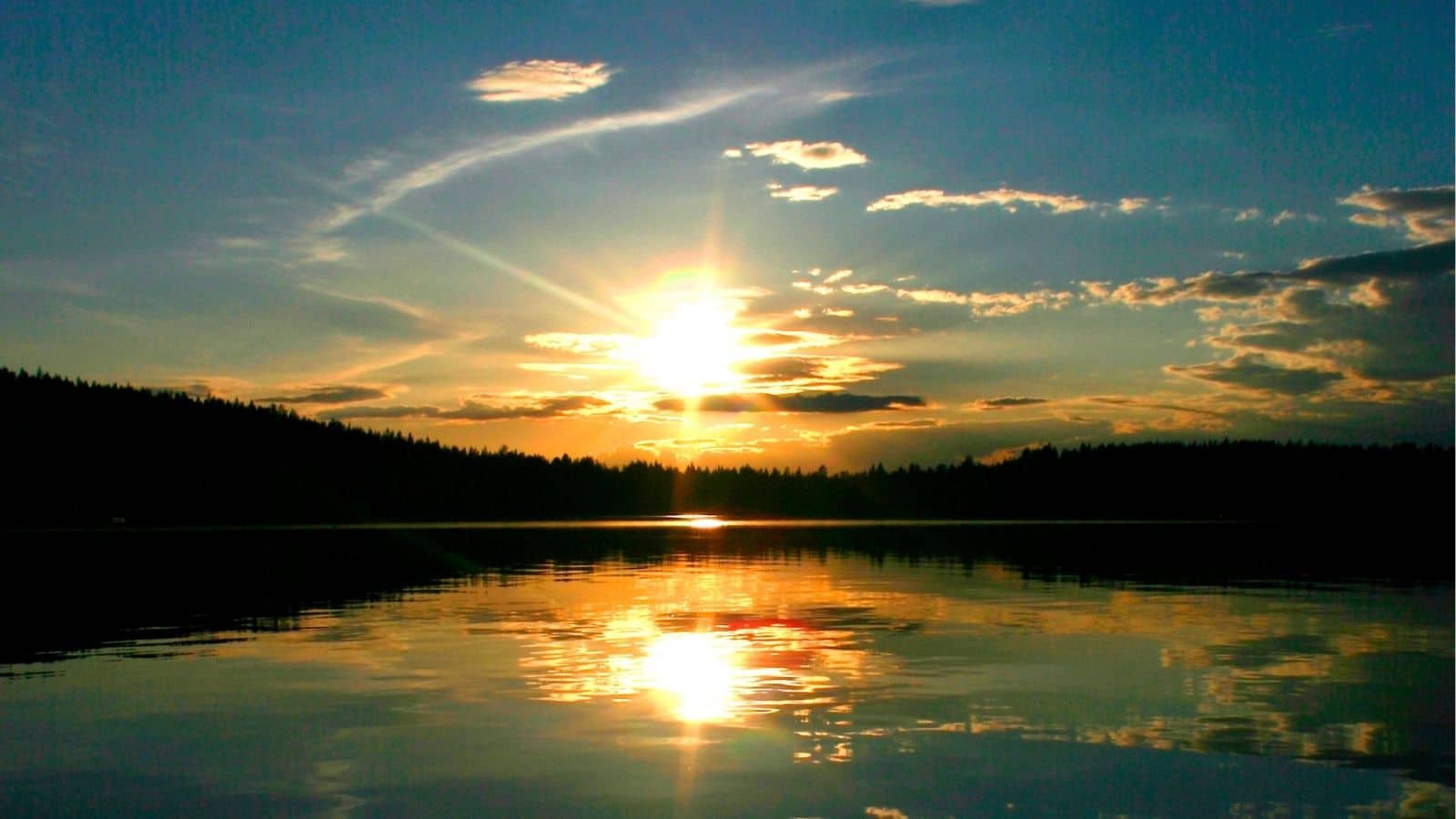 The sun sets over a calm lake, casting reflections of the sky and clouds on the water, with a tree-lined horizon in the background.