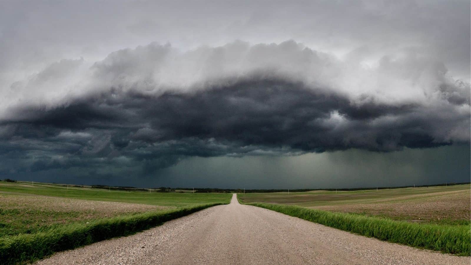 A gravel road stretches into the distance under a dramatic, dark storm cloud, with fields of grass and crops on either side.