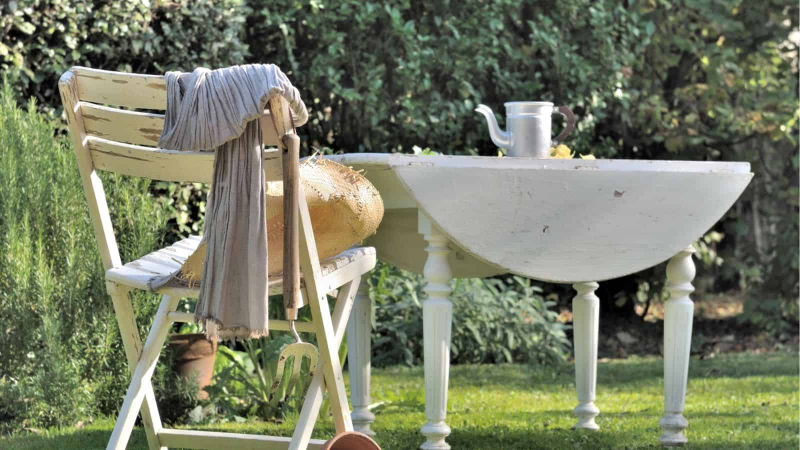 A weathered white chair and table sit on grass in a garden; a straw hat and cloth rest on the chair, and a metal teapot is on the table.