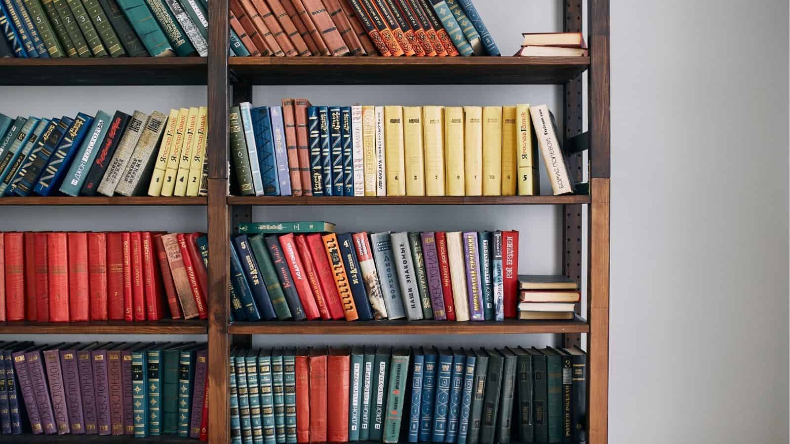 A bookshelf filled with books arranged by color, including blue, green, yellow, and red volumes, with a few books stacked horizontally on the top right shelf.