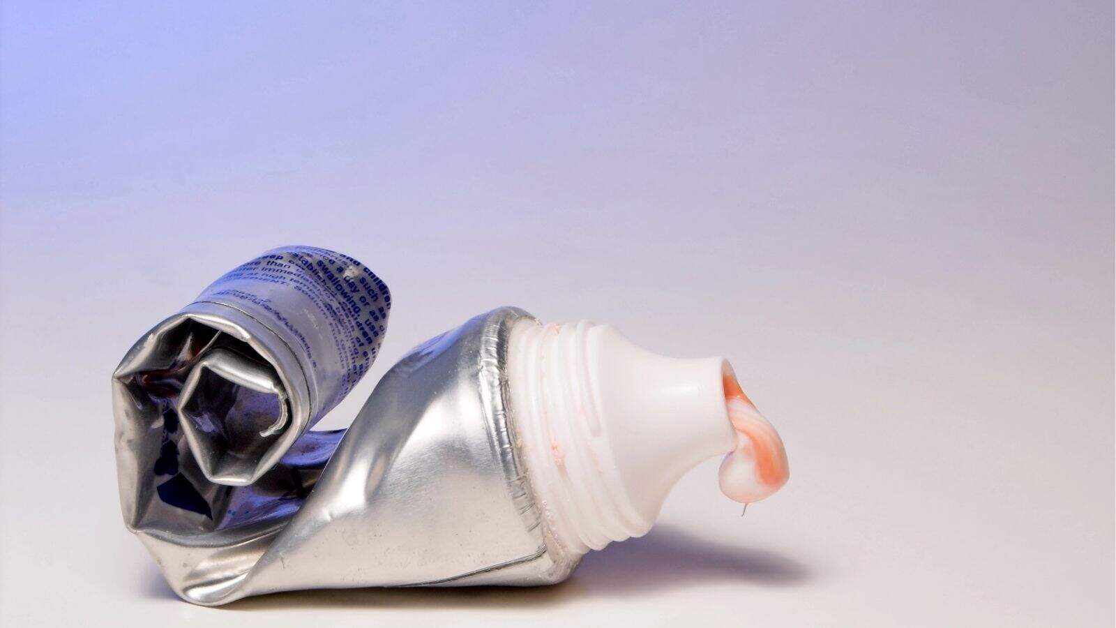 A squeezed metallic toothpaste tube with its cap off, showing pink and white striped toothpaste coming out of the opening, against a plain background.