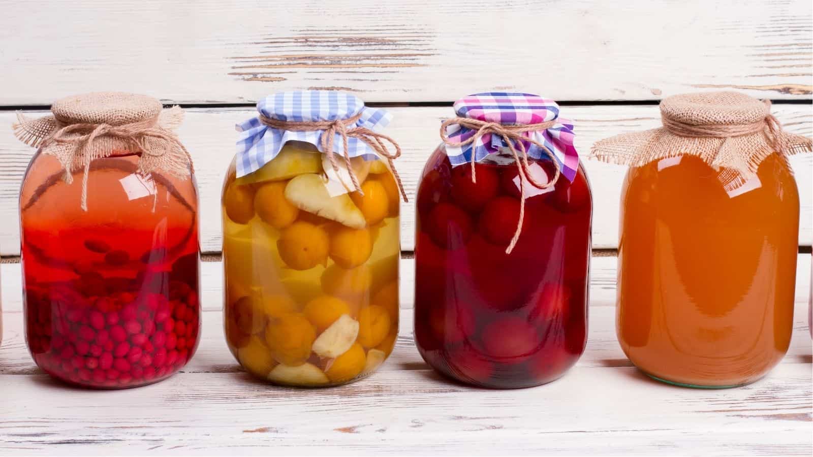 Four glass jars filled with preserved fruits and liquids, each covered with a fabric lid, are lined up in front of a white wooden background.