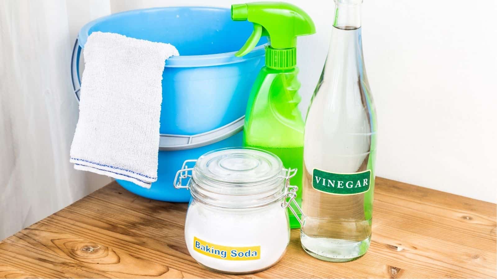 A jar labeled “Baking Soda,” a bottle of vinegar, a green spray bottle, a blue bucket with a white cloth, all on a wooden surface.