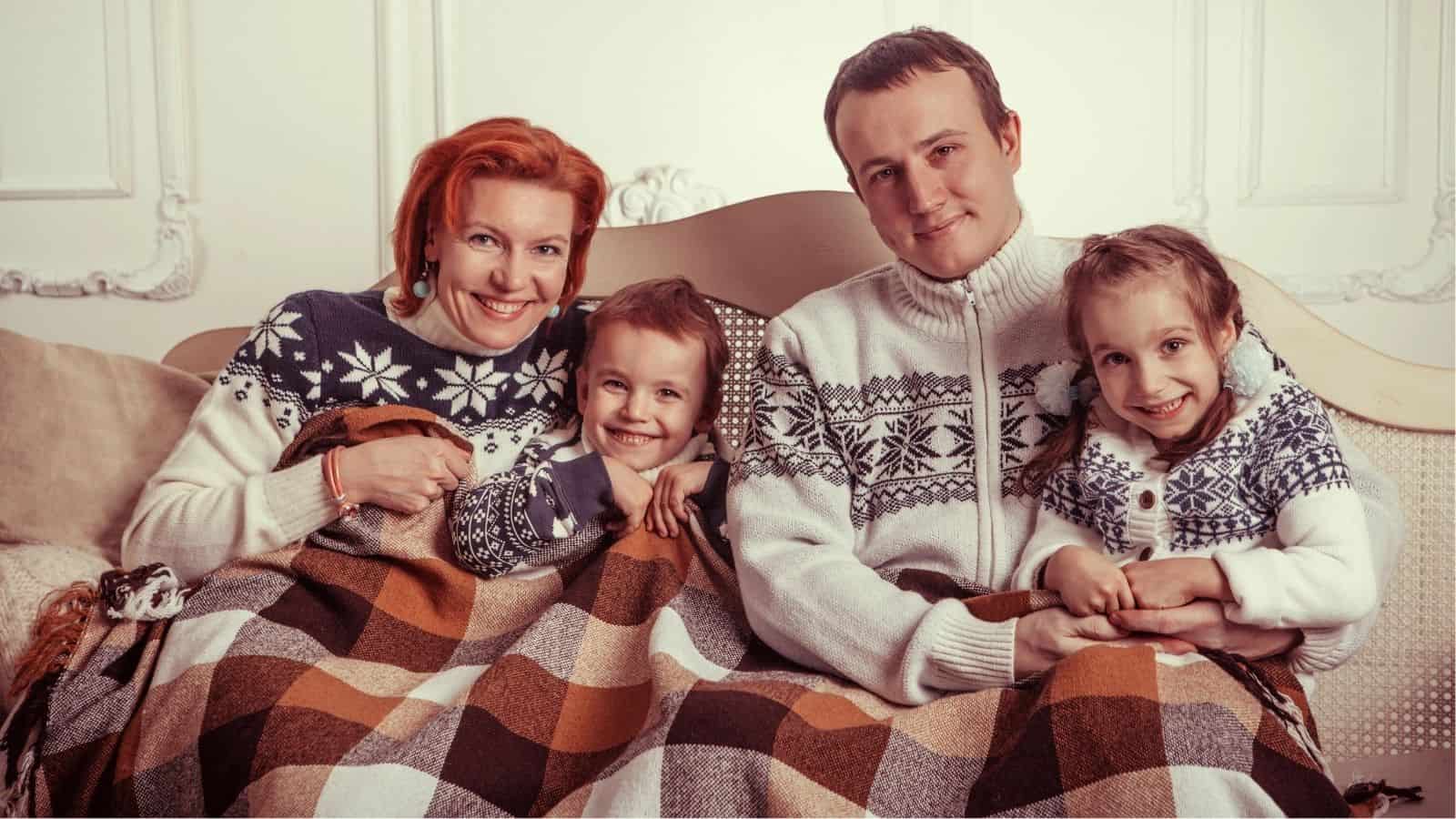 A family of four wearing winter sweaters sit on a couch, smiling and wrapped together in a brown plaid blanket.