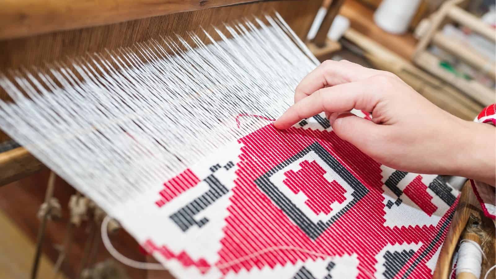 A person weaves a red, white, and black geometric patterned textile on a loom, guiding threads by hand.
