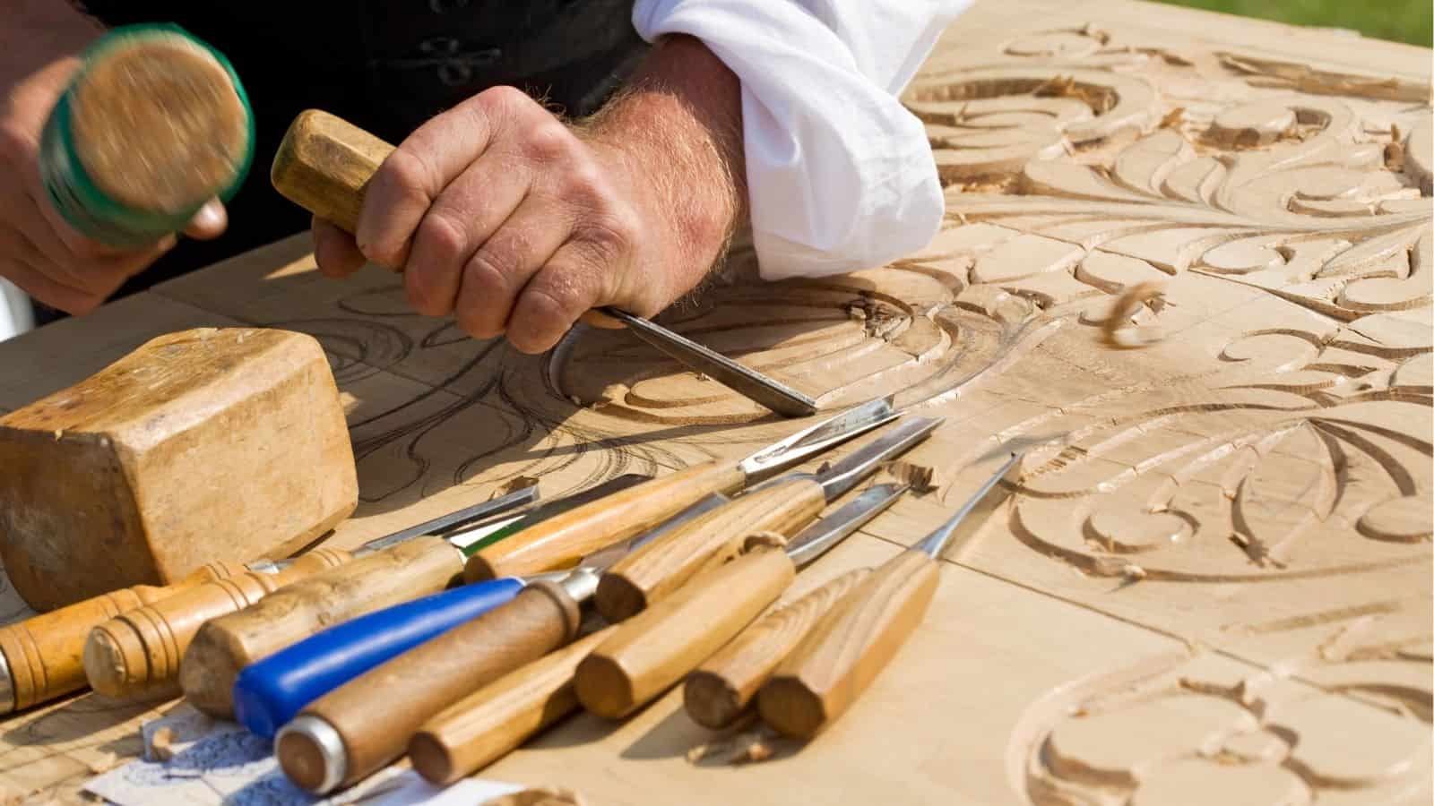 A person uses a chisel and mallet to carve detailed patterns into a wooden surface, with various woodworking tools lying nearby.