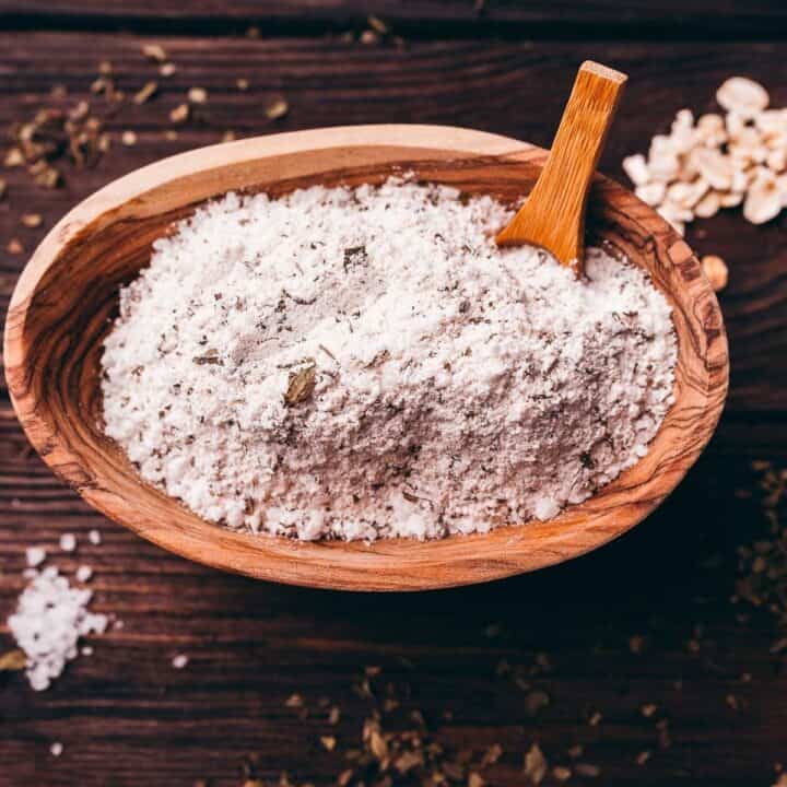A wooden bowl filled with a white, powdery flour sits on a wooden surface, with a small wooden scoop resting inside the bowl.