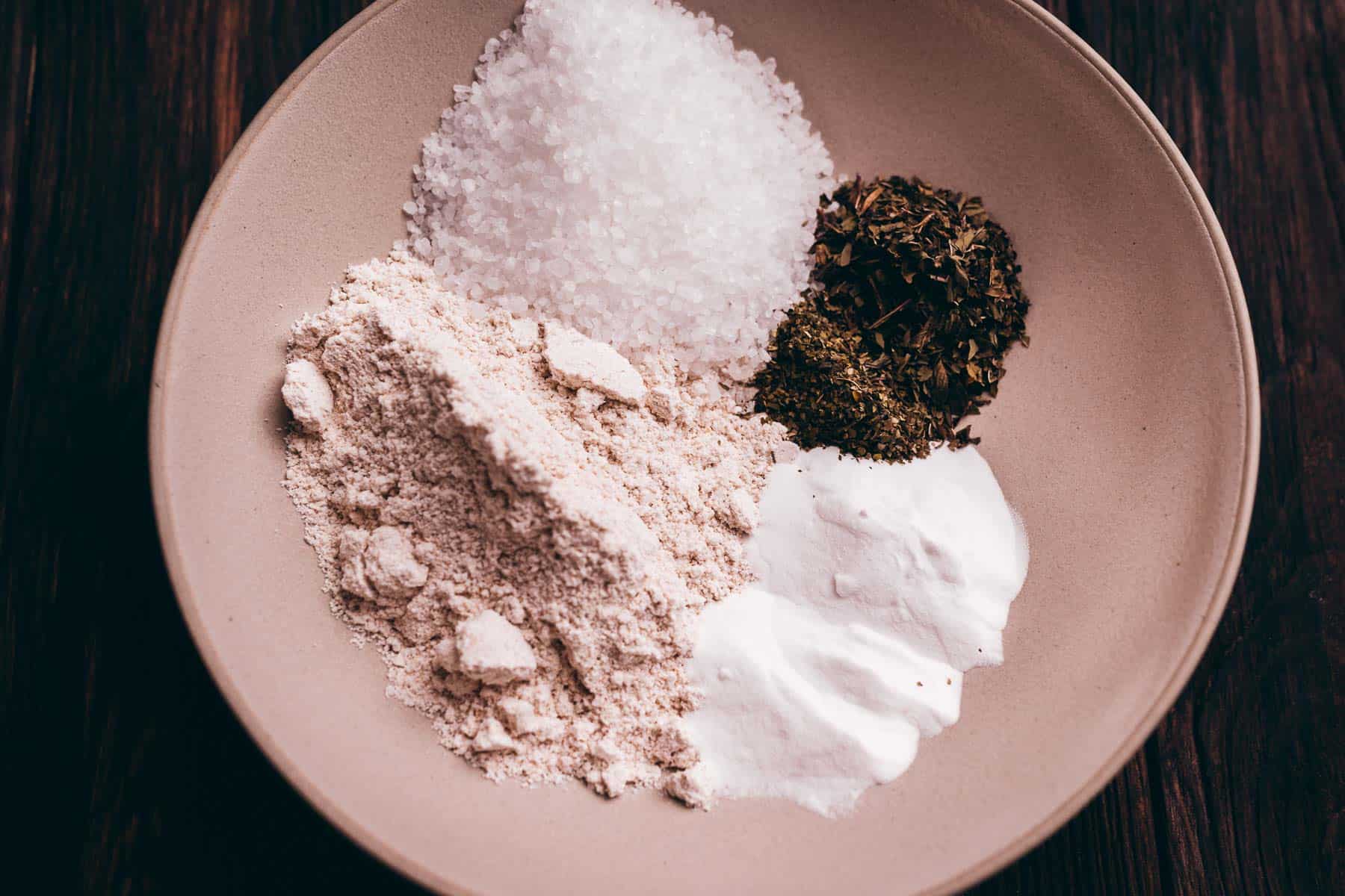 A beige bowl containing sea salt, dried herbs, flour, and baking powder, each ingredient arranged in separate small piles on a dark wooden surface.