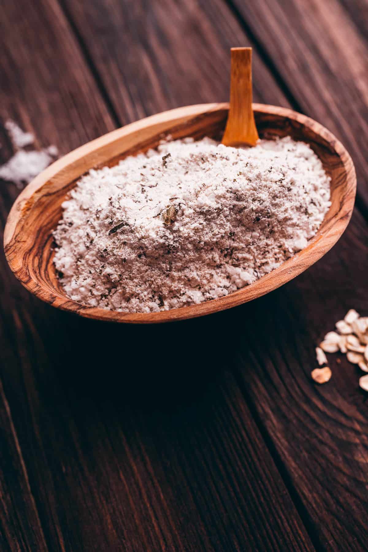 A wooden bowl filled with oat flour and a small wooden scoop, placed on a dark wooden surface with some loose oats nearby.