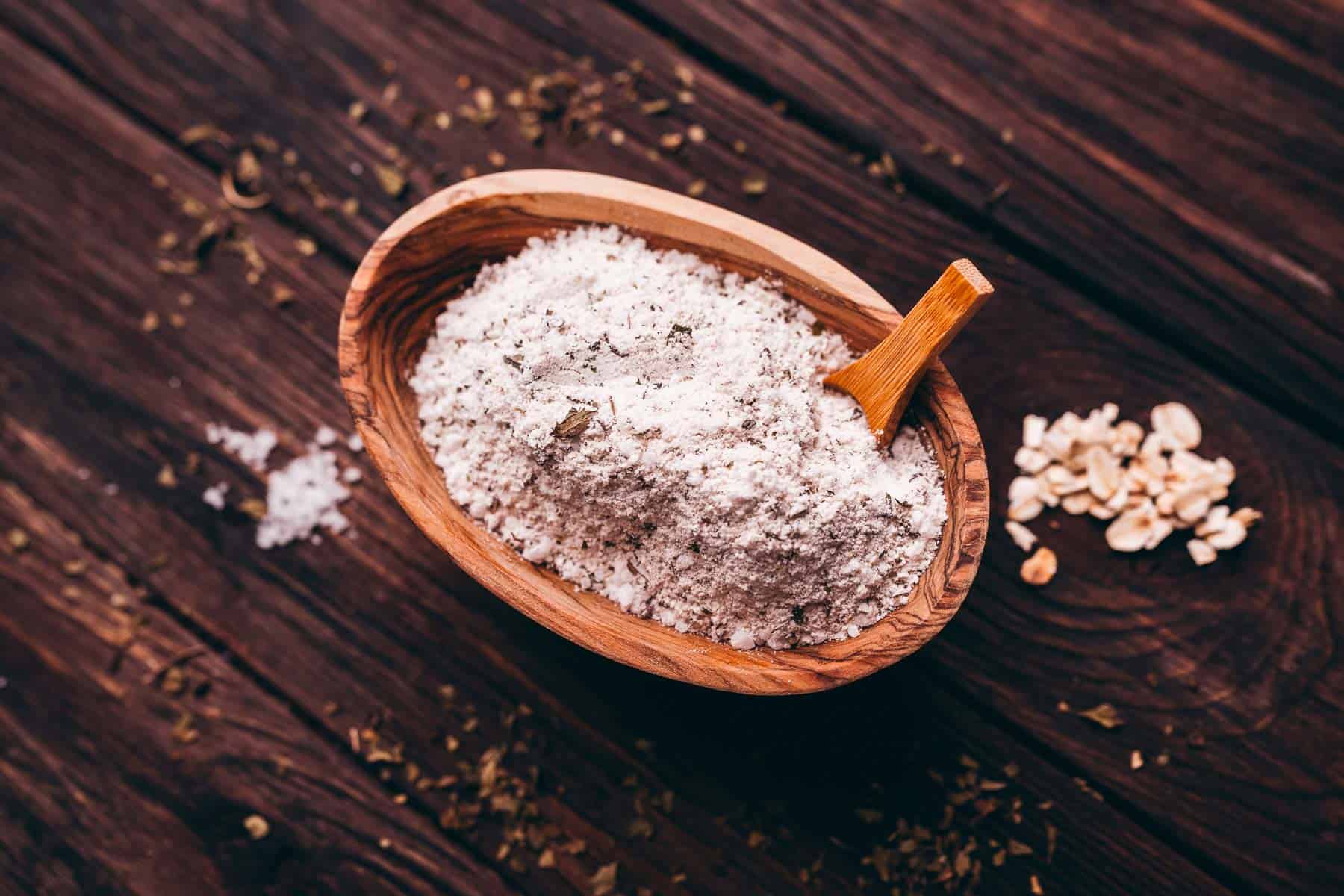 Wooden bowl containing white powder, possibly flour or ground oats, with a wooden spoon on a rustic wooden surface. Some scattered oats and herbs are nearby.