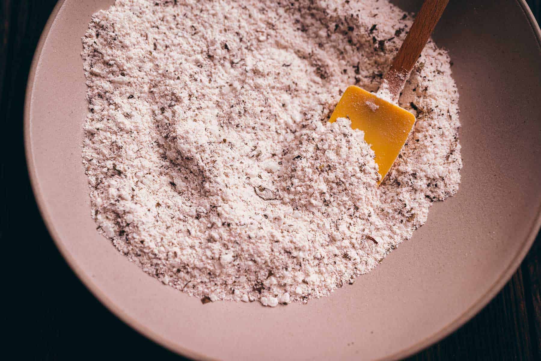 A ceramic bowl containing a dry mixture of flour and herbs, with a yellow spatula resting inside.