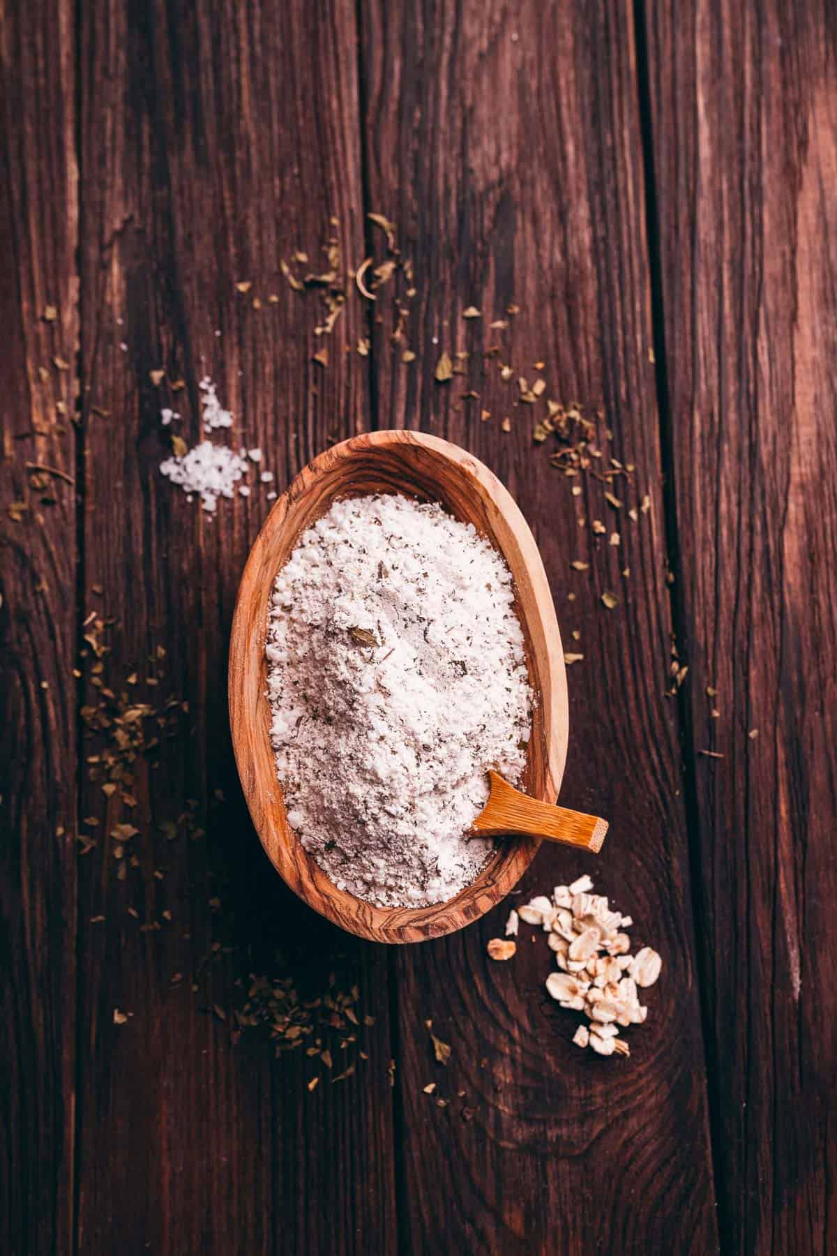A wooden bowl filled with flour and a small wooden scoop sits on a dark wooden surface, with oats and herbs scattered nearby.