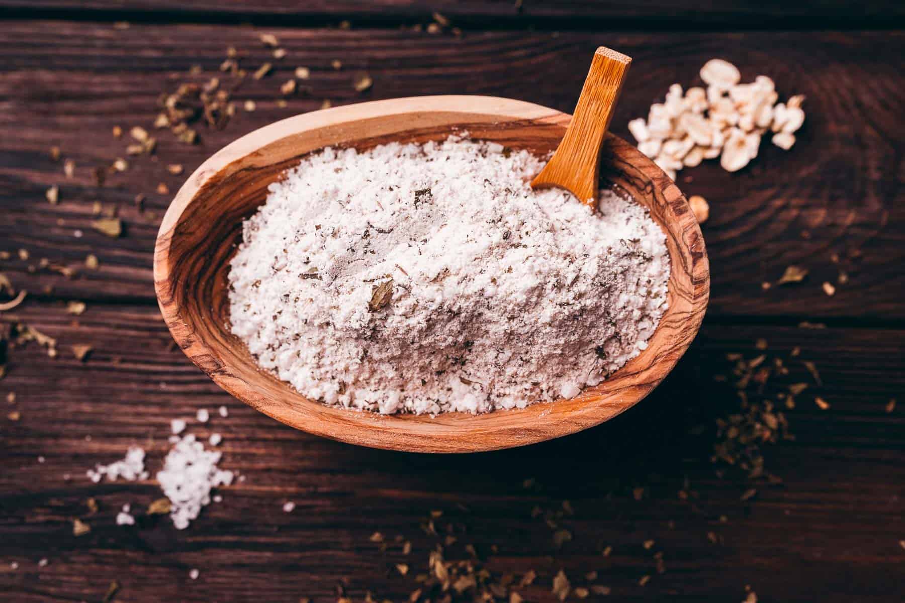 A wooden bowl filled with flour and a wooden scoop sits on a dark wooden surface, with some flour and herbs scattered nearby.