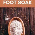 A wooden bowl filled with a powdered foot soak mix sits on a wooden surface with scattered dried herbs and oats.