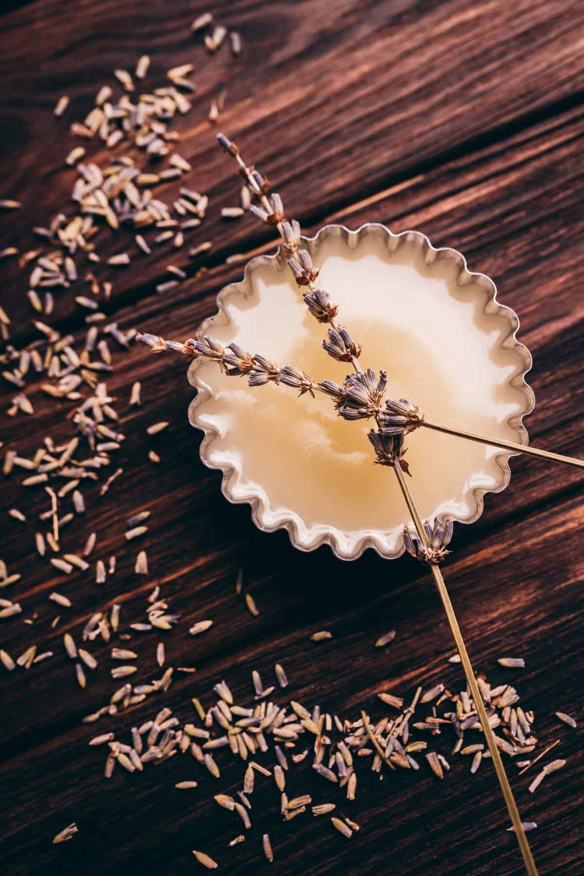 A small fluted tin filled with solid balm, topped with dried lavender sprigs, sits on a wooden surface scattered with loose lavender buds.