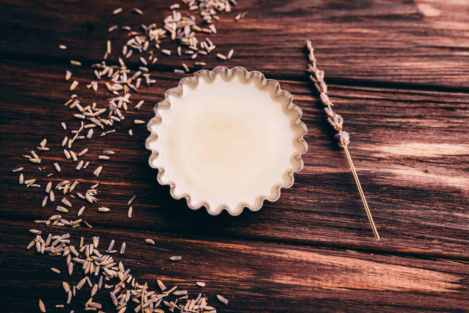 A fluted metal dish filled with solid wax sits on a wooden surface, surrounded by scattered dried lavender and a single lavender sprig.