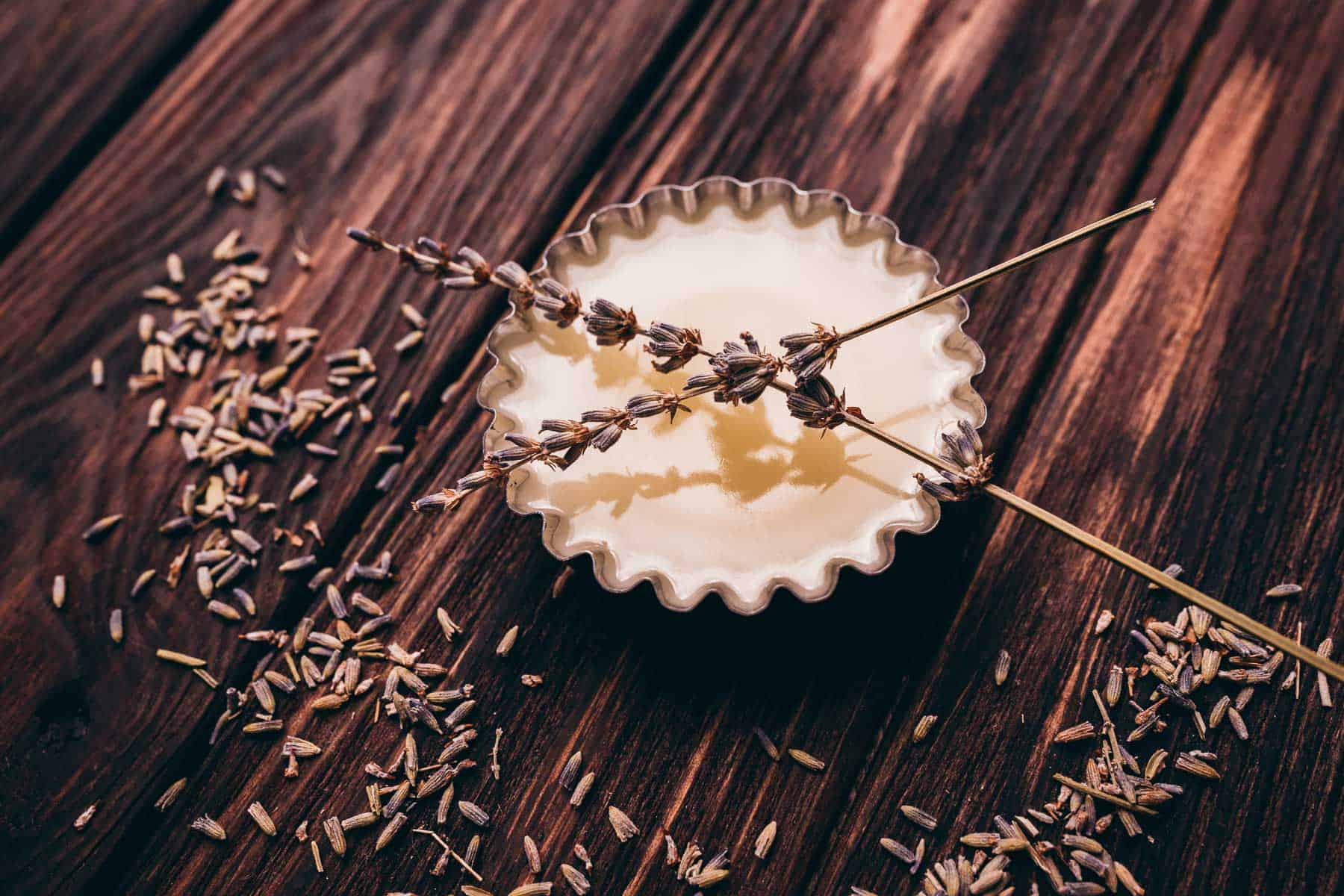 A small metal tin filled with creamy balm, topped with two sprigs of dried lavender, sits on a wooden surface scattered with loose lavender buds.