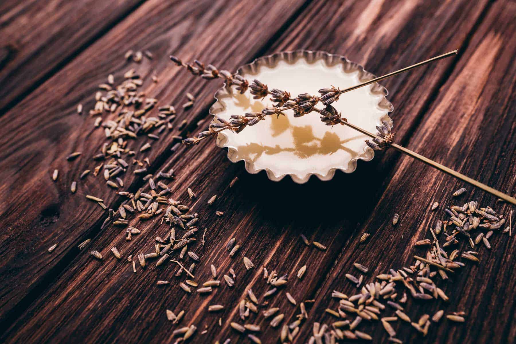 Small dish of pale yellow balm with two sprigs of dried lavender on top, surrounded by scattered lavender buds on a wooden surface.