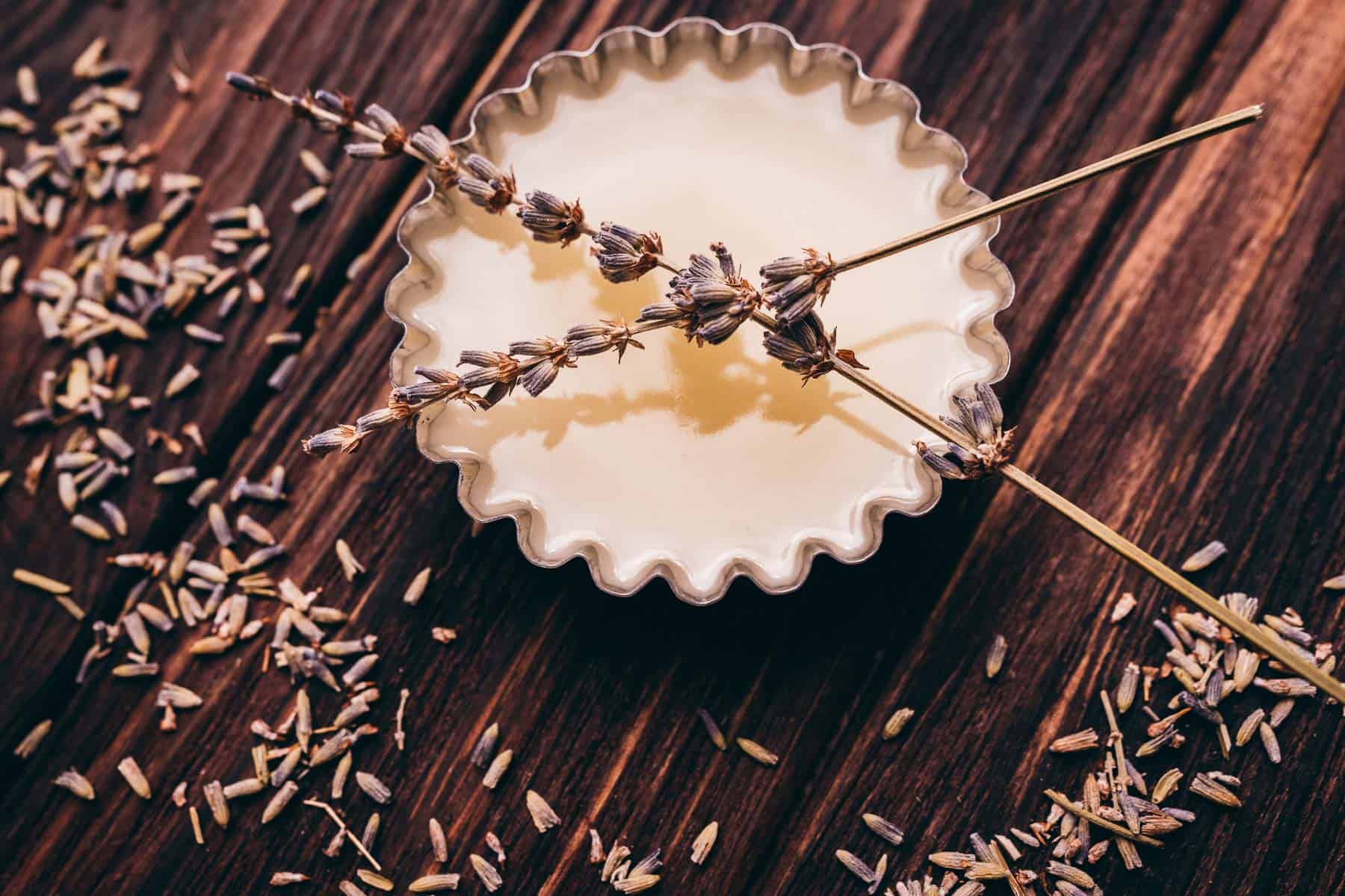 A white fluted dish filled with liquid and topped with dried lavender sprigs, surrounded by scattered lavender buds on a wooden surface.