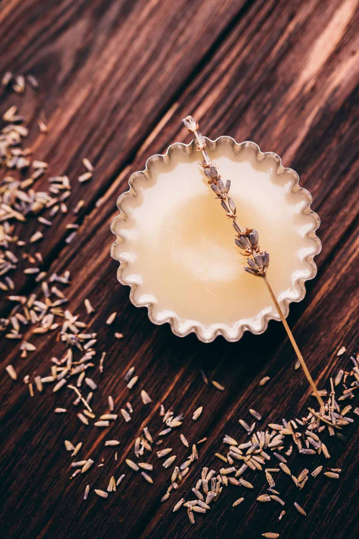 A small metal tin of solid balm with a sprig of dried lavender on top, surrounded by scattered lavender buds on a wooden surface.