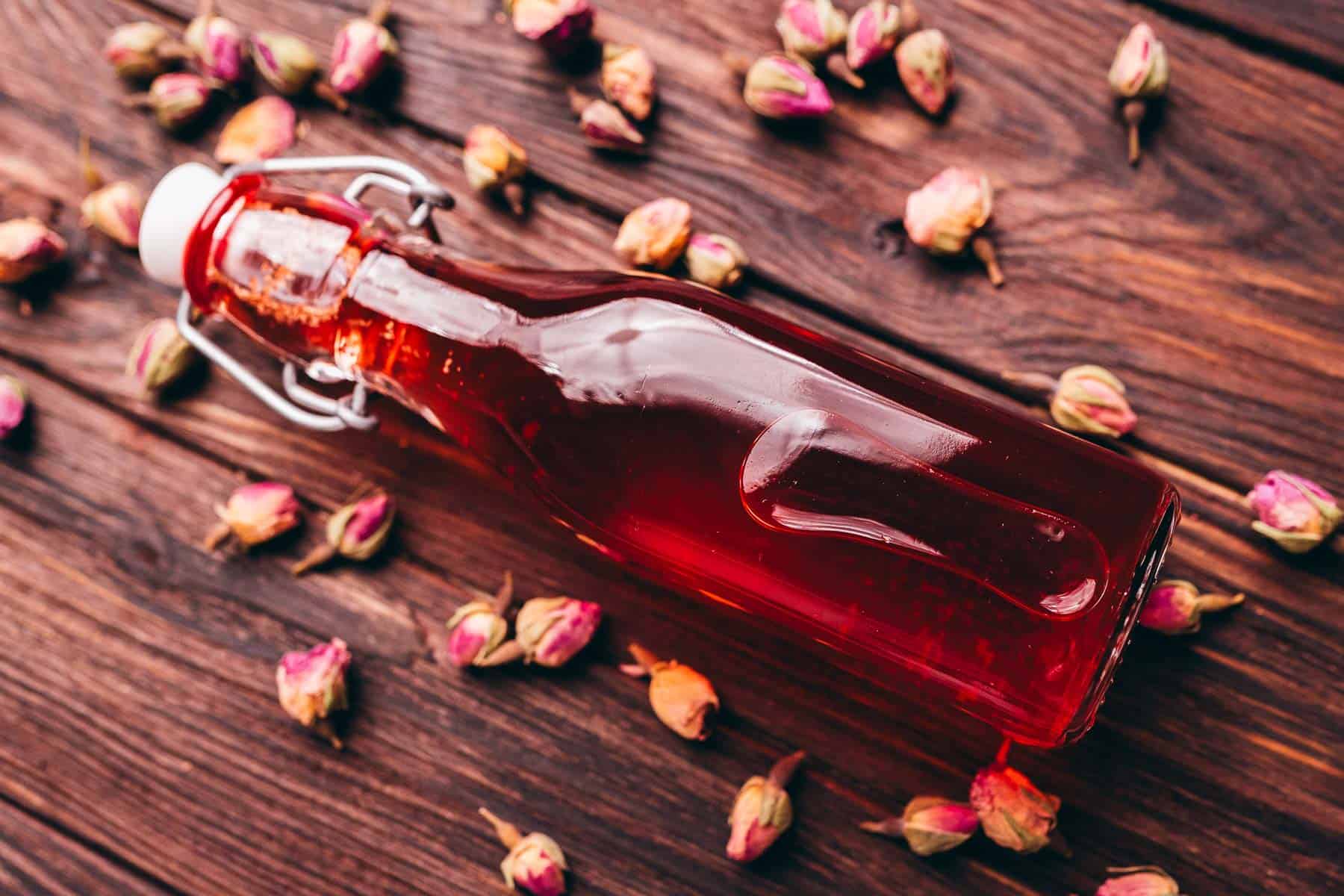 A glass bottle filled with red rose vinegar lies on a wooden surface, surrounded by scattered dried rosebuds.