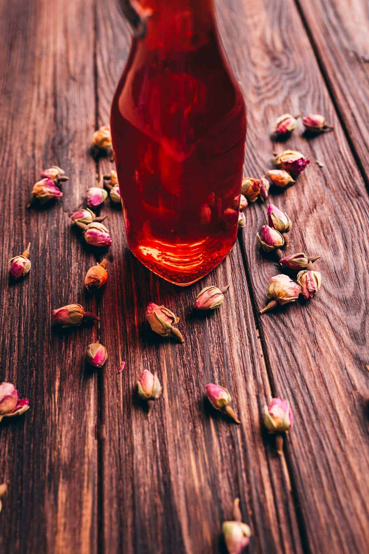 A glass bottle filled with rose vinegar stands on a wooden surface, surrounded by scattered dried rosebuds.
