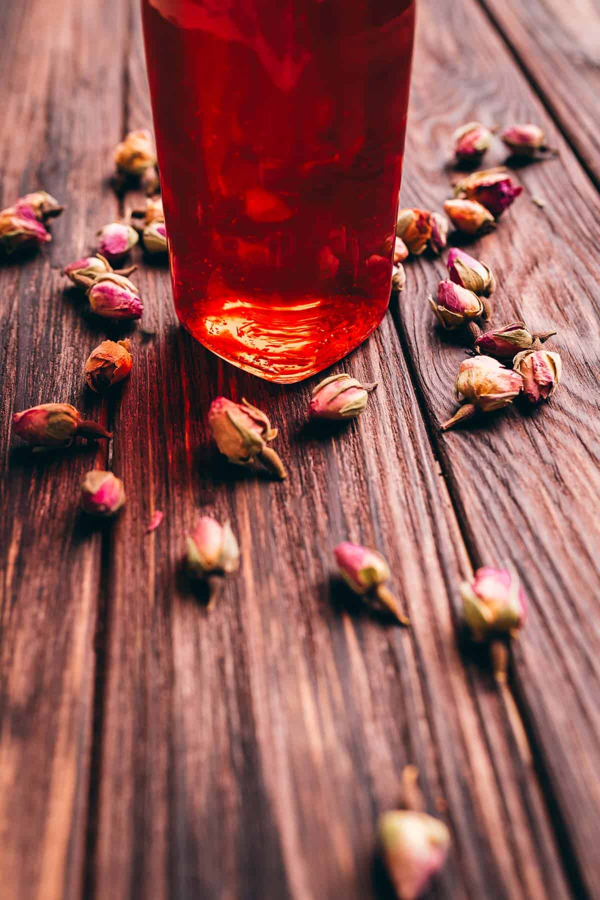 A glass of red beverage infused with rose vinegar sits on a wooden table, surrounded by scattered dried rosebuds.