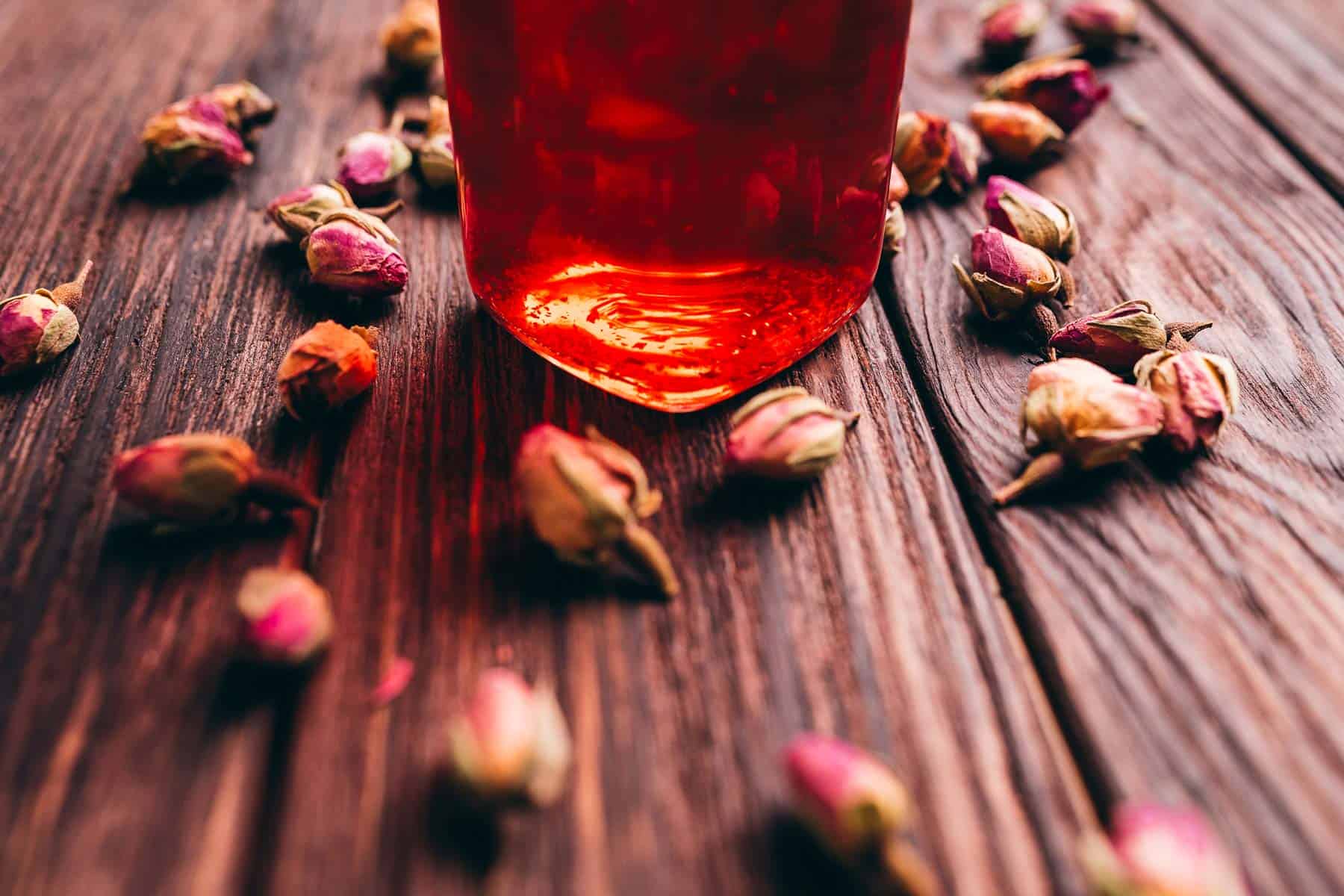 A glass jar filled with amber-colored rose vinegar sits on a wooden surface, surrounded by scattered dried rosebuds.