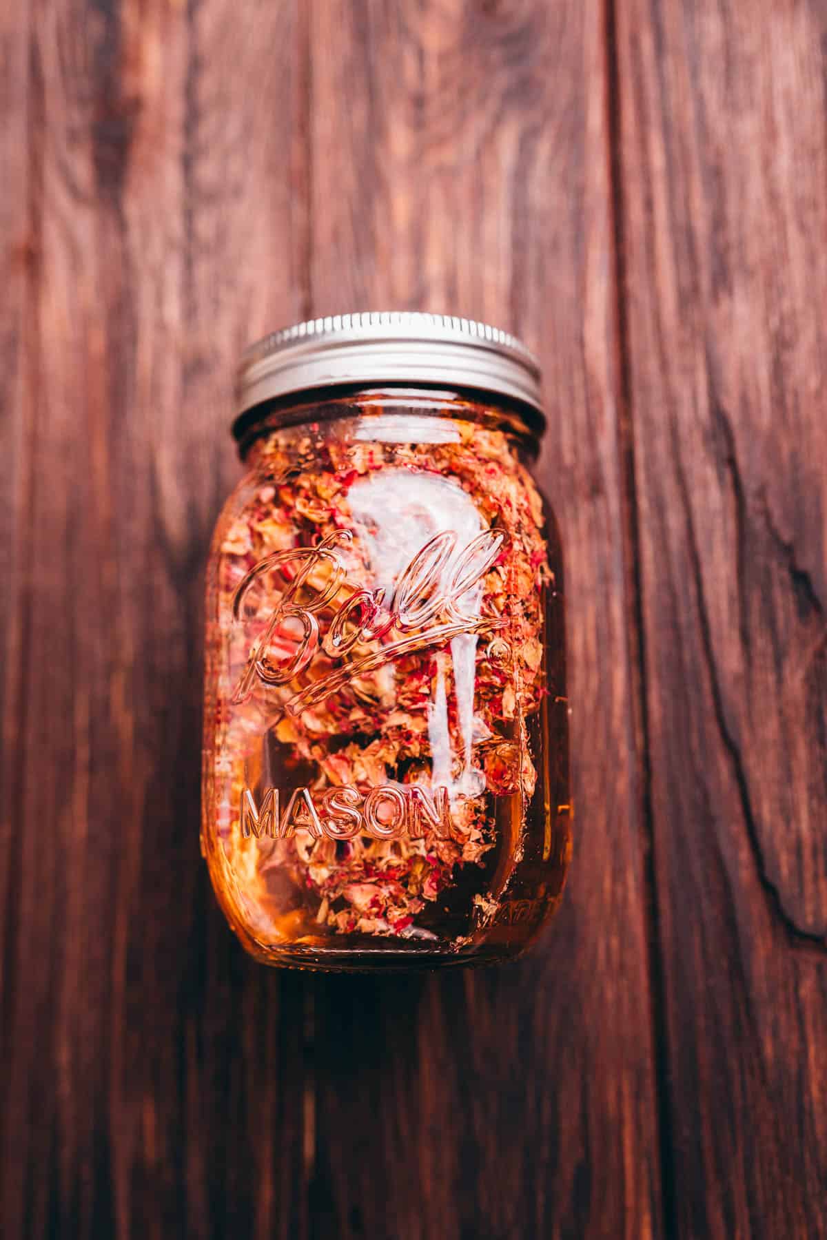 A clear Ball mason jar filled with dried rose petals and rose vinegar sits on a wooden surface.