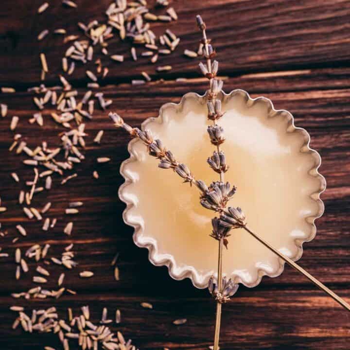A tin of creamy balm with dried lavender sprigs on top, surrounded by scattered lavender buds on a wooden surface.
