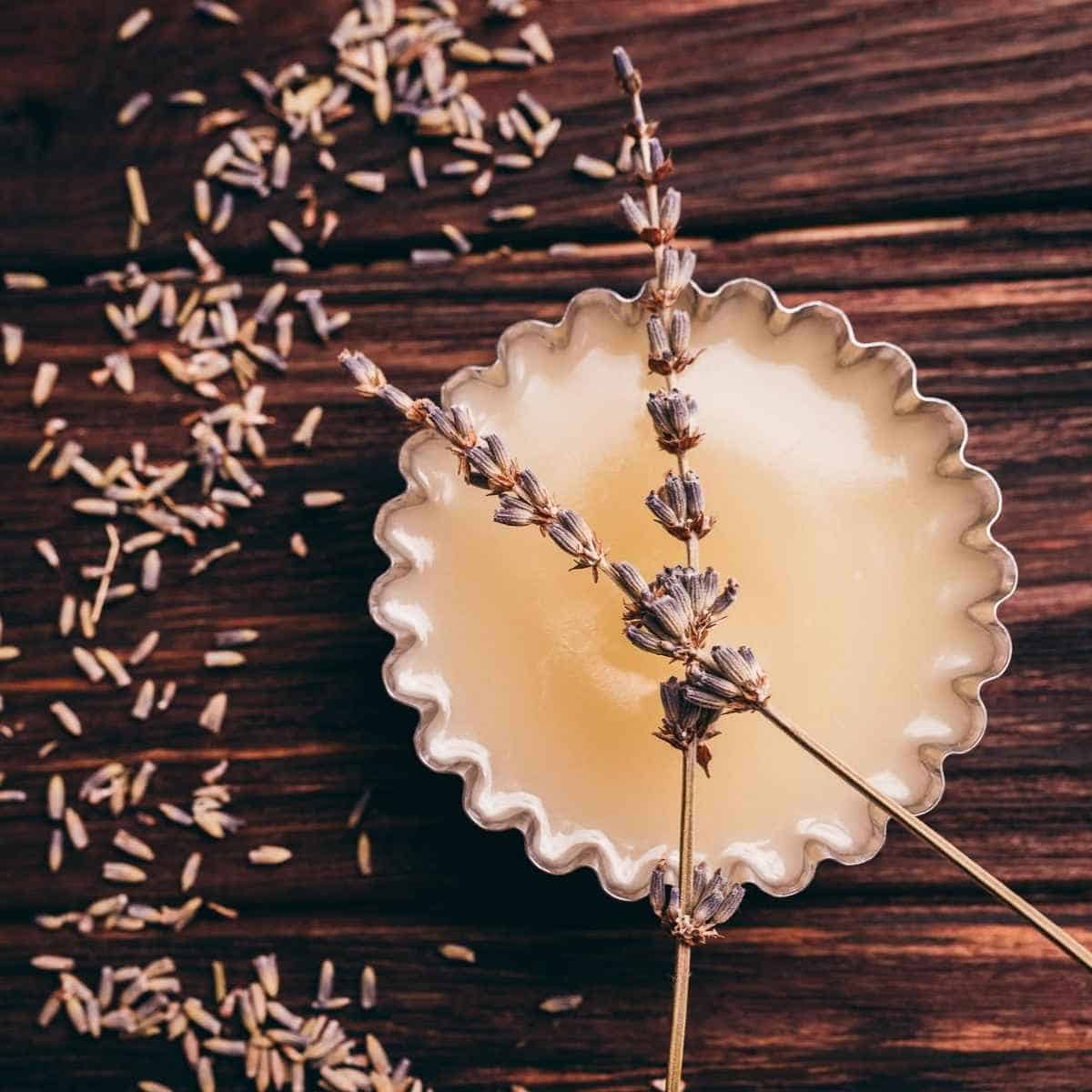 A tin of creamy balm with dried lavender sprigs on top, surrounded by scattered lavender buds on a wooden surface.